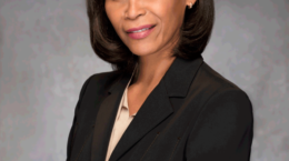 Pamela Newkirk, with straight, shoulder-length brown hair, wears a black blazer over a beige top. She stands and smiles softly against a plain gray background at FreedomCon 2022.