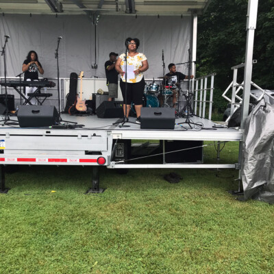A band performs on an outdoor stage at the 2021 Festival in Sewickley PA, with a keyboardist, guitarist, drummer, and singer in front. The stage is set up on grass with trees behind and musical equipment visible for Juneteenth Day. A band performs on an outdoor stage at the 2021 Festival in Sewickley PA, with a keyboardist, guitarist, drummer, and singer in front. The stage is set up on grass with trees behind and musical equipment visible for Juneteenth Day.