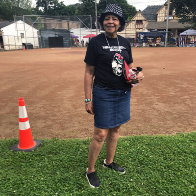 A woman stands on grass near a baseball field at the 2021 Festival in Sewickley, PA, wearing a black t-shirt, denim skirt, sneakers, and a polka-dot hat. She holds sunglasses and a red item, smiling by an orange traffic cone. Trees and buildings are behind her. A woman stands on grass near a baseball field at the 2021 Festival in Sewickley, PA, wearing a black t-shirt, denim skirt, sneakers, and a polka-dot hat. She holds sunglasses and a red item, smiling by an orange traffic cone. Trees and buildings are behind her.