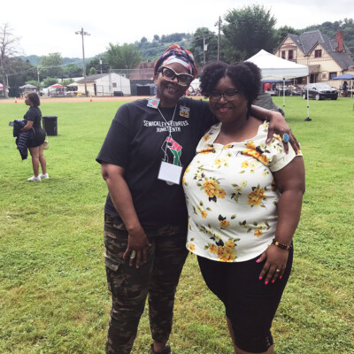 Two women smile and pose together outdoors at the 2021 Festival in Sewickley, PA. One wears a headwrap, black shirt, and camo pants; the other sports a white floral top and black shorts. Tents and people fill the Juneteenth Day celebration behind them. Two women smile and pose together outdoors at the 2021 Festival in Sewickley, PA. One wears a headwrap, black shirt, and camo pants; the other sports a white floral top and black shorts. Tents and people fill the Juneteenth Day celebration behind them.