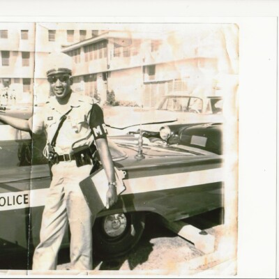 A smiling police officer in uniform stands beside a vintage police car, holding a clipboard. The scene, possibly from a Memphis Area Branch ASALH monthly meeting, features mid-20th century buildings and parked cars in the background. A smiling police officer in uniform stands beside a vintage police car, holding a clipboard. The scene, possibly from a Memphis Area Branch ASALH monthly meeting, features mid-20th century buildings and parked cars in the background.