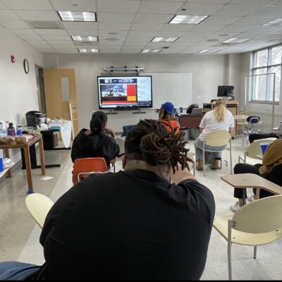 A group of people sit spaced apart in a classroom, watching a Dr. Charles R. Drew Knowledge Bowl presentation on a large screen. Some chairs are empty; snacks and drinks are on a side table. The room has large windows and fluorescent lighting. A group of people sit spaced apart in a classroom, watching a Dr. Charles R. Drew Knowledge Bowl presentation on a large screen. Some chairs are empty; snacks and drinks are on a side table. The room has large windows and fluorescent lighting.