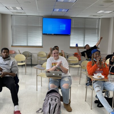 A group of students sit in a classroom with desks and backpacks, some smiling and clapping, while others hold drinks and food. The Dr. Charles R. Drew Knowledge Bowl is shown on a large screen behind them. A group of students sit in a classroom with desks and backpacks, some smiling and clapping, while others hold drinks and food. The Dr. Charles R. Drew Knowledge Bowl is shown on a large screen behind them.