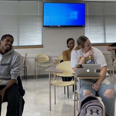 A group of students sits in a classroom with desks arranged in rows, enjoying drinks or snacks. Some are smiling or laughing during the Knowledge Bowl. A student uses a laptop, while a monitor and windows are visible in the background. A group of students sits in a classroom with desks arranged in rows, enjoying drinks or snacks. Some are smiling or laughing during the Knowledge Bowl. A student uses a laptop, while a monitor and windows are visible in the background.