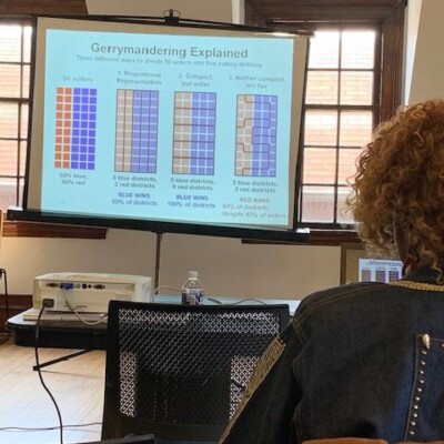 A presentation slide titled Gerrymandering Explained shows three diagrams comparing proportional, compact, and neither compact nor fair districting methods with colored boxes representing voters and illustrating how election outcomes can impact African Americans. Audience members are seated in front of the screen. A presentation slide titled Gerrymandering Explained shows three diagrams comparing proportional, compact, and neither compact nor fair districting methods with colored boxes representing voters and illustrating how election outcomes can impact African Americans. Audience members are seated in front of the screen.