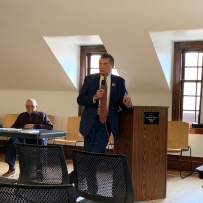 A man in a suit speaks at a podium about the importance of the vote and upcoming election in a bright room with wooden floors and large windows, while another man sits behind him. Several empty chairs and a projector screen are visible. A man in a suit speaks at a podium about the importance of the vote and upcoming election in a bright room with wooden floors and large windows, while another man sits behind him. Several empty chairs and a projector screen are visible.