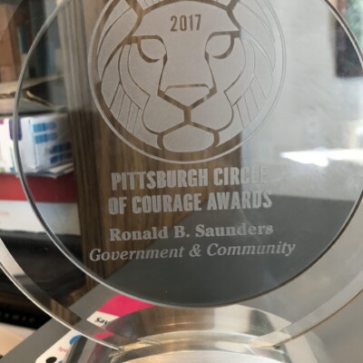 A glass award engraved with a lion’s face, the text “2017 Pittsburgh Circle of Courage Awards, Ronald B. Saunders, Government & Community,” and a McKenzie ASALH Branch Award card advertising a free trial is placed beneath the base. A glass award engraved with a lion’s face, the text “2017 Pittsburgh Circle of Courage Awards, Ronald B. Saunders, Government & Community,” and a McKenzie ASALH Branch Award card advertising a free trial is placed beneath the base.