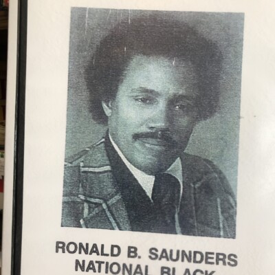 A printed poster features a black-and-white portrait of a man in a suit. The text reads: FROM COMMUNITY SERVICE at the top and RONALD B. SAUNDERS NATIONAL BLACK POLITICAL CAUCUS at the bottom, highlighting the Ronald B. Saunders Awards. A printed poster features a black-and-white portrait of a man in a suit. The text reads: FROM COMMUNITY SERVICE at the top and RONALD B. SAUNDERS NATIONAL BLACK POLITICAL CAUCUS at the bottom, highlighting the Ronald B. Saunders Awards.